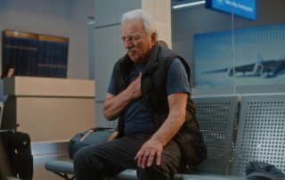 Elderly man sitting in medical lobby, painfully holding his chest.